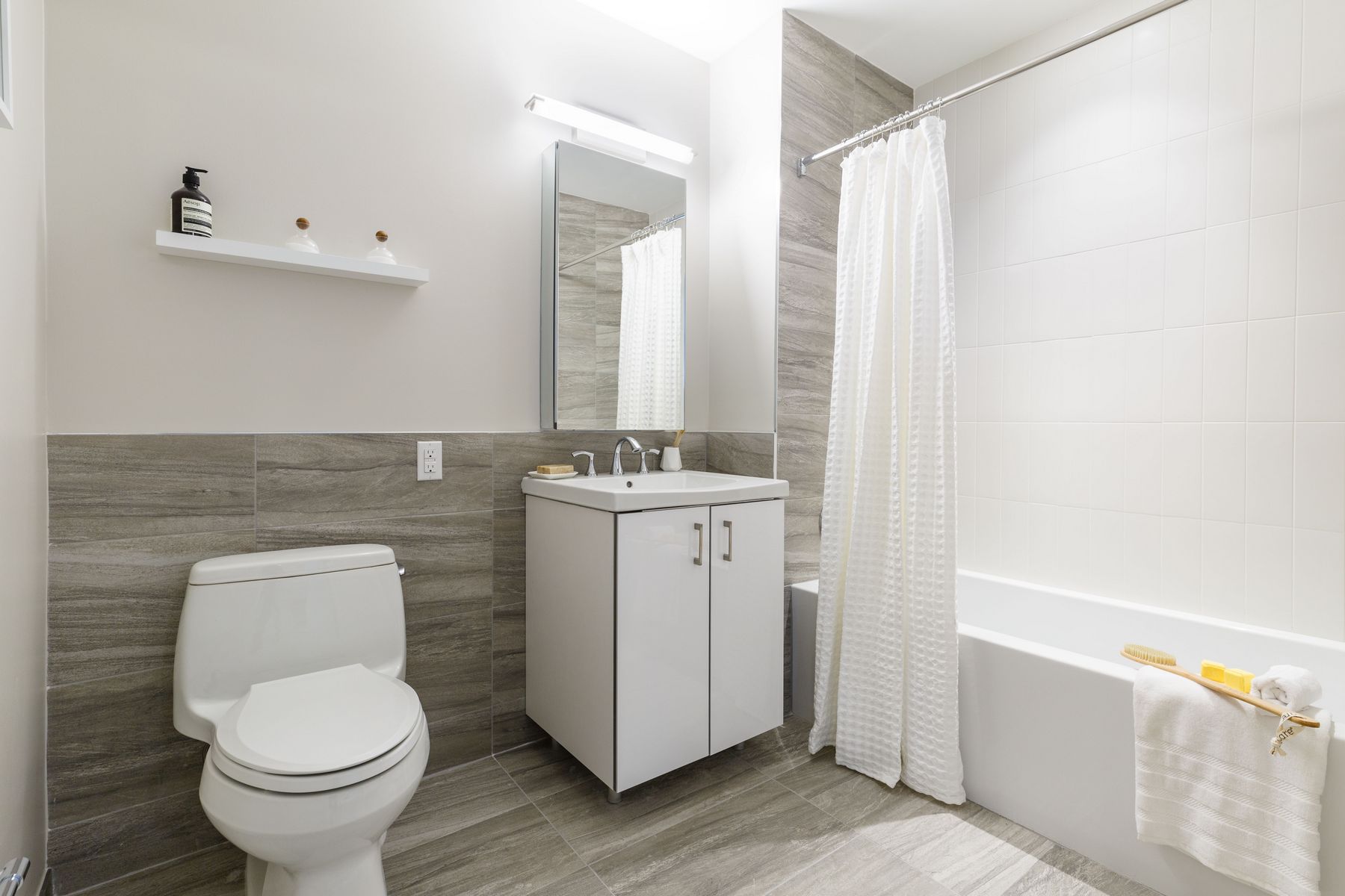 Modern bathroom with white tub shower combo, fully tiled in white and grayish marble type, single vanity with sink and mirror, large tiles on floor and partially up wall, single white shelf above white toilet